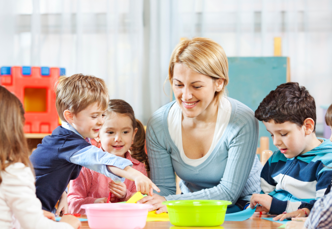 Afbeelding van juf die met 4 kinderen aan een tafel zit met 2 bakjes en gekleurd papier